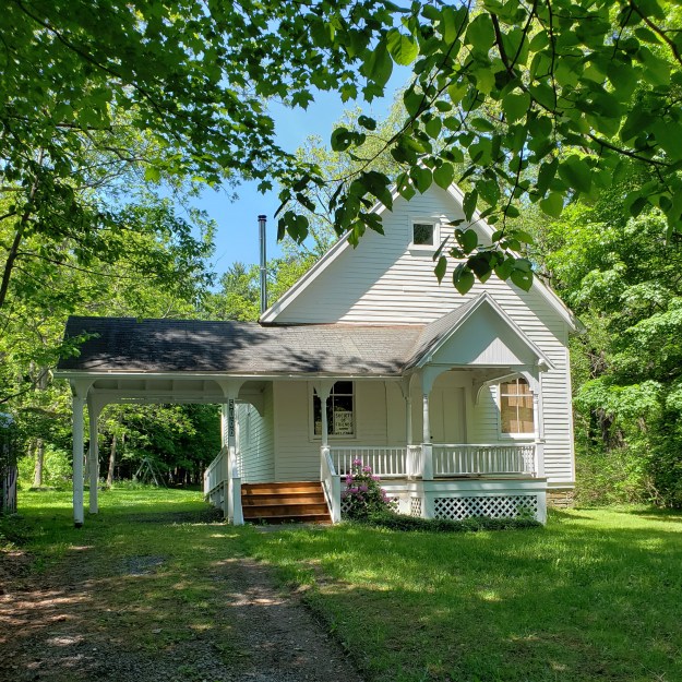 A small, white house with a portico on the left side, surrounded by trees and bright green grass